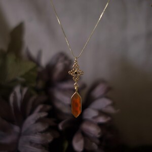Op de afbeelding: Een goudkleurige ketting met een hanger met een oranjerode edelsteen. De hanger hangt aan een fijne ketting en heeft een sierlijke bovenkant. De achtergrond is wazig, met donkerpaarse bloemen. Sieraad.