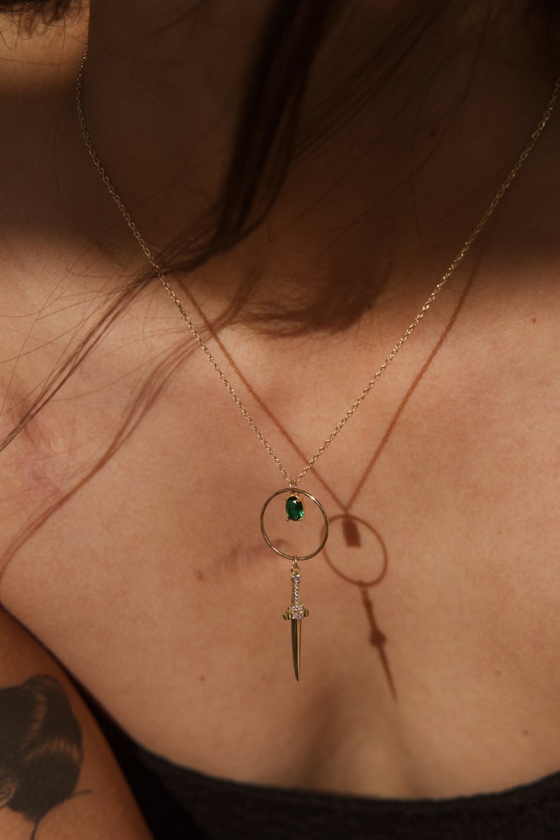 May include: A gold necklace with a green gemstone pendant and a sword charm. The sword charm is adorned with small clear gemstones.
