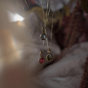May include: Three delicate gold necklaces featuring oval-shaped pendants. One pendant contains a light blue stone, another a deep pink stone, and the third a dark green stone. Each pendant is suspended from a thin gold chain with small accents.