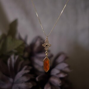Op de afbeelding: Een goudkleurige ketting met een hanger met een oranje, geometrische edelsteen. De hanger hangt aan een fijne ketting en heeft een sierlijke, decoratieve bovenkant met een kleine zwarte steen. De achtergrond is vervaagd met donkerpaarse bloemen.