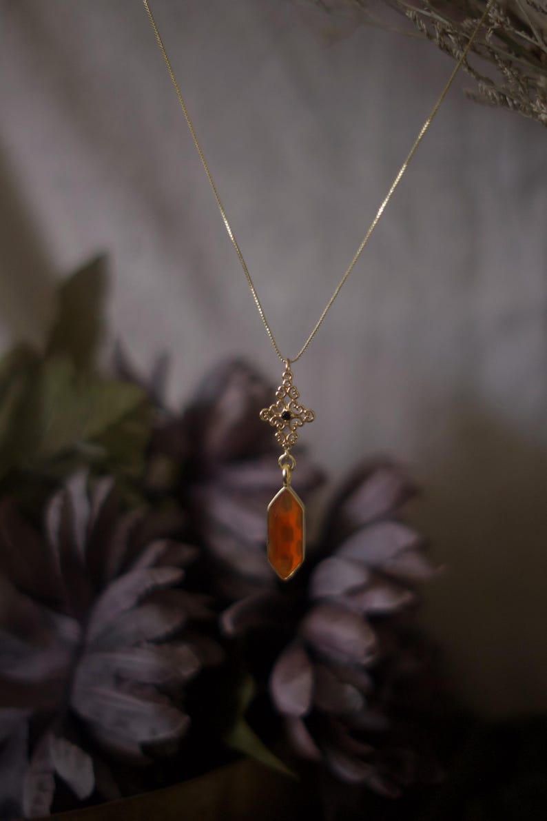Op de afbeelding: Een goudkleurige ketting met een hanger met een oranje, geometrische edelsteen. De hanger hangt aan een fijne ketting en heeft een sierlijke, decoratieve bovenkant met een kleine zwarte steen. De achtergrond is vervaagd met donkerpaarse bloemen.