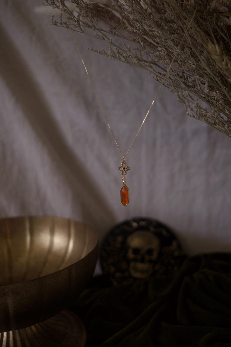 Op de afbeelding: Een goudkleurige ketting met een hanger. De hanger heeft een kruisontwerp met zwarte accenten en een druppelvormige oranje edelsteen. De ketting hangt tegen een neutrale achtergrond met gedroogde bloemen en een decoratieve kom.