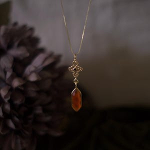 Op de afbeelding: Een goudkleurige ketting met een fijne ketting. De hanger heeft een sierlijk, opengewerkt ontwerp met een rood-oranje, facetgeslepen edelsteen. De ketting wordt getoond tegen een vage, donkere achtergrond, waardoor de details van de sieraden worden benadrukt.