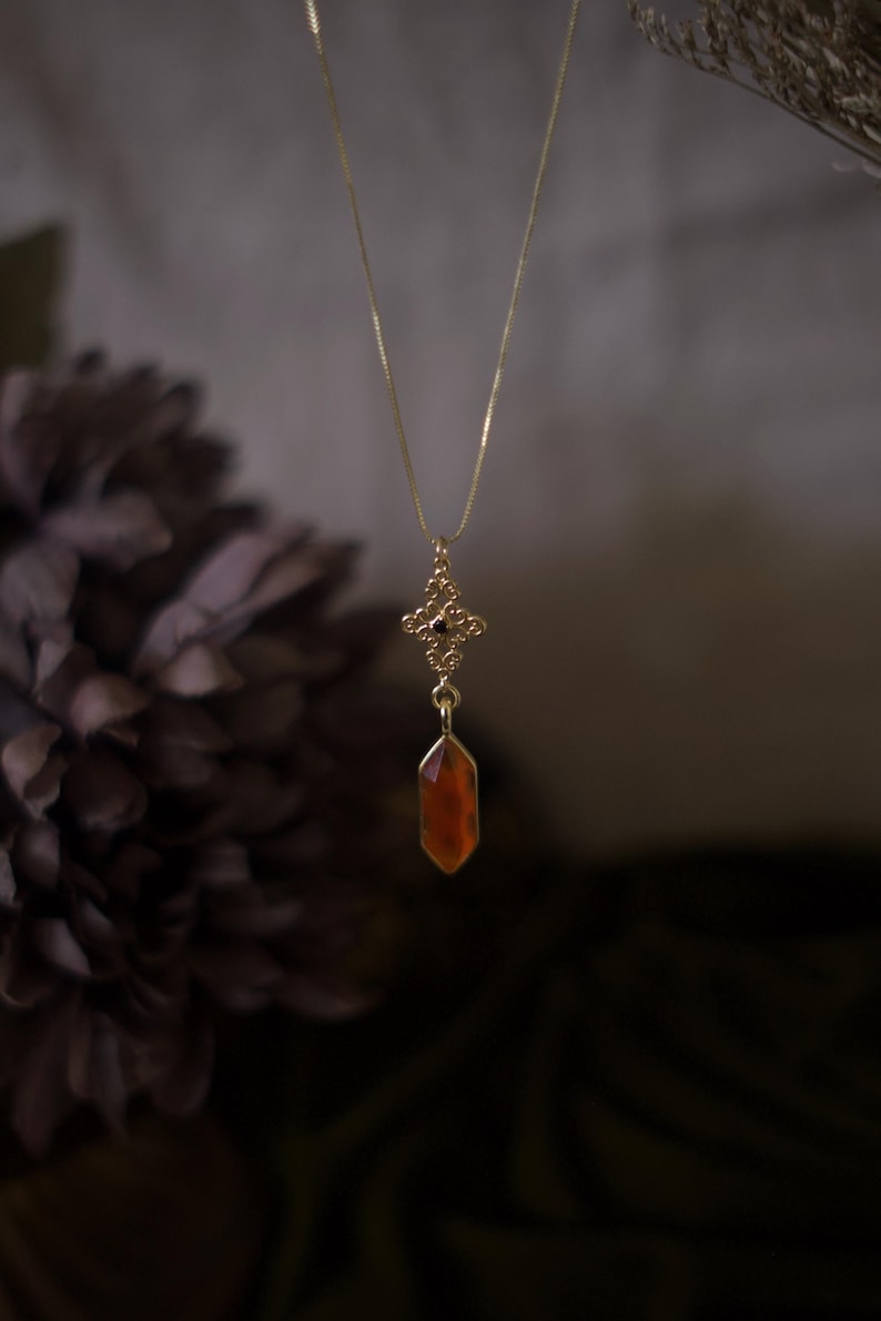Op de afbeelding: Een goudkleurige ketting met een fijne ketting. De hanger heeft een sierlijk, opengewerkt ontwerp met een rood-oranje, facetgeslepen edelsteen. De ketting wordt getoond tegen een vage, donkere achtergrond, waardoor de details van de sieraden worden benadrukt.