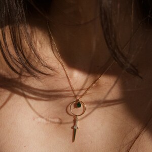 May include: A gold chain necklace with a green gemstone pendant and a small sword charm.