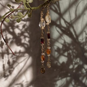 Puede incluir: Pendientes colgantes dorados con una variedad de piedras preciosas, incluyendo cuentas claras, rojas y &aacute;mbar. Los pendientes cuelgan de una delicada cadena y se exhiben sobre un fondo de una rama frondosa y sombras.