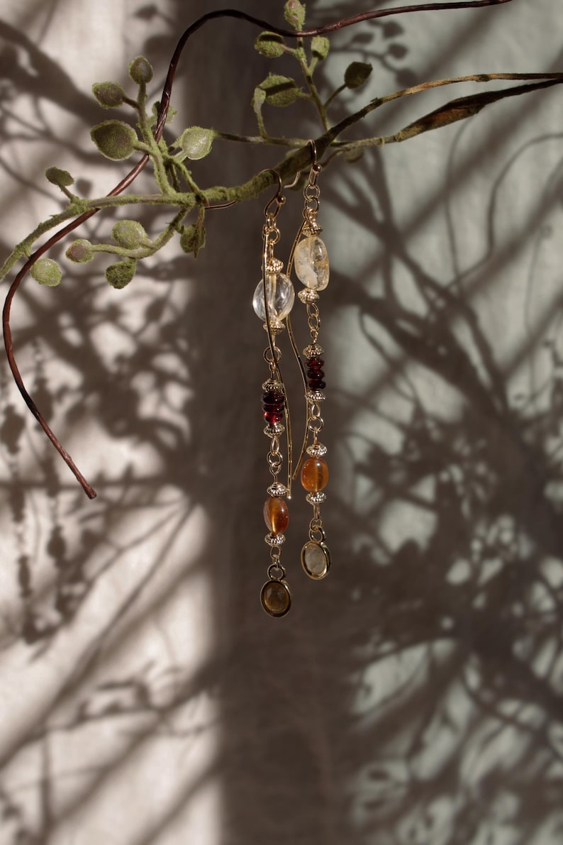 Puede incluir: Pendientes colgantes dorados con una variedad de piedras preciosas, incluyendo cuentas claras, rojas y &aacute;mbar. Los pendientes cuelgan de una delicada cadena y se exhiben sobre un fondo de una rama frondosa y sombras.