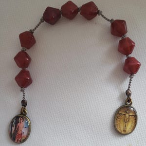 Puede incluir: Un rosario de piedras preciosas rojas con una cadena plateada y dos colgantes ovalados. Un colgante presenta una representación de un santo, y el otro presenta una representación de Jesús en la cruz.
