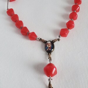May include: A red glass rosary with a silver chain and two oval-shaped medallions featuring religious imagery. The medallions have a gold-coloured frame.