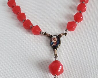 Op de afbeelding: Een rode glazen rozenkrans met een zilveren ketting en twee ovale medaillons met religieuze afbeeldingen. De medaillons hebben een gouden rand.