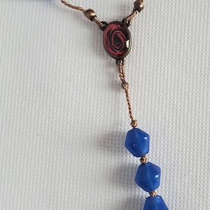 May include: A blue glass rosary with a brown cord and a metal charm featuring a rose and an angel.