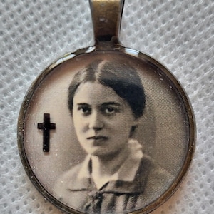 Puede incluir: Un colgante de estilo vintage con un retrato en tonos sepia de una mujer. El colgante redondo tiene un marco de color bronce y una pequeña cruz negra en el lado izquierdo. La fianza también es de color bronce.