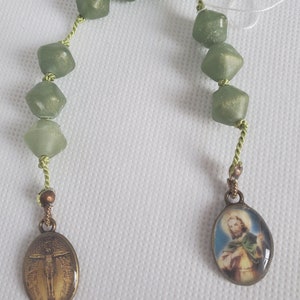 Puede incluir: Un rosario de cuentas verdes con una medalla de santo y una medalla de crucifijo. El rosario está hecho de pequeñas cuentas facetadas y está enhebrado en un cordón verde.