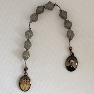 Puede incluir: Rosario de cuentas de vidrio gris con un colgante de metal marrón que presenta una imagen de un hombre. El colgante en el otro extremo del rosario presenta una imagen de un hombre en una cruz.