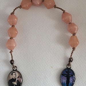 Puede incluir: Un rosario de cuentas rosadas con un cordón marrón y dos dijes de metal. Un dije presenta una imagen de un hombre con un traje negro y el otro dije presenta una cruz con un diseño de vitral.
