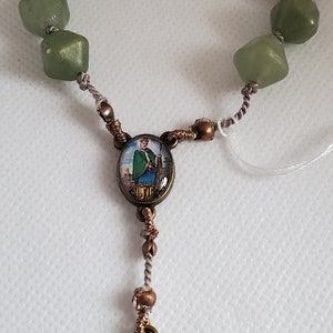 May include: A green beaded rosary with a brown metal pendant featuring a Celtic cross and a smaller pendant with a picture of a saint.