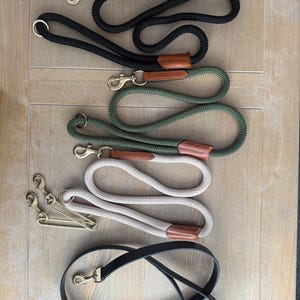 Könnte beinhalten: Eine Sammlung von vier Hundeleinen, bestehend aus drei geflochtenen Seilleinen in Schwarz, Olivgrün und Beige mit hellbraunen Lederakzenten und Messingbeschlägen sowie einer glatten, flachen schwarzen Lederleine mit einem Messingkarabiner.