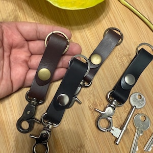 Puede incluir: Llaveros de cuero en marrón y negro con herrajes metálicos. Los llaveros tienen un cierre a presión y un cierre giratorio. También se ven varias llaves.