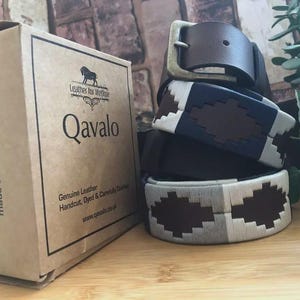 Puede incluir: Una pila de cinturones de cuero con costuras decorativas en marrón, blanco y azul marino. Una caja de cartón marrón con la marca "Qavalo" y el texto "Leather accessories made to last a lifetime" es visible.