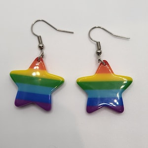 Puede incluir: Un par de pendientes en forma de estrella con un esquema de colores arcoíris. Cada pendiente presenta una estrella de cinco puntas con rayas horizontales en rojo, naranja, amarillo, verde, azul y morado. Los pendientes tienen ganchos plateados.