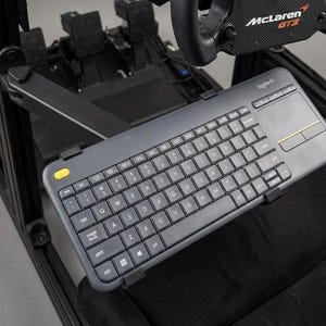 Könnte beinhalten: Eine graue Logitech-Funk-Tastatur mit Touchpad ist an einem schwarzen Haltearm befestigt. Die Tastatur hat schwarze Tasten und eine gelbe Taste. Das Bild zeigt auch ein Lenkrad und Pedale, was auf ein Rennsimulator-Setup hindeutet.
