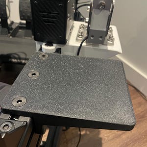 Op de afbeelding: Een zwart getextureerd platform met afgeronde hoeken, bevestigd aan een race-simulatoropstelling. Het platform is vastgezet met drie bouten. Andere componenten zijn een zwarte doos met koolstofvezeltextuur en een zilverkleurig metalen onderdeel.