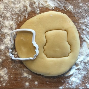 Könnte beinhalten: Ein weißer Plastik-Ausstecher in Form eines Zylinders, neben einem runden Stück Keksteig, in dem die Form des Zylinders ausgeschnitten ist.