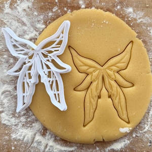 May include: A white plastic butterfly-shaped cookie cutter next to a round piece of dough with a butterfly imprint. The dough is on a wooden cutting board dusted with flour. The cookie cutter is a kitchen tool for baking.