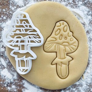 Puede incluir: Un cortador de galletas blanco con forma de hongo junto a una huella con forma de hongo en la masa. La masa es de color marrón claro y está espolvoreada con harina blanca. El cortador de galletas tiene un diseño detallado.