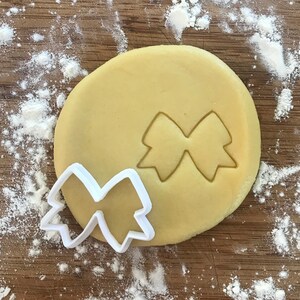 Pode incluir: Um cortador de biscoitos de pl&aacute;stico branco em forma de la&ccedil;o, em cima de uma pe&ccedil;a redonda de massa de biscoito n&atilde;o cozida.