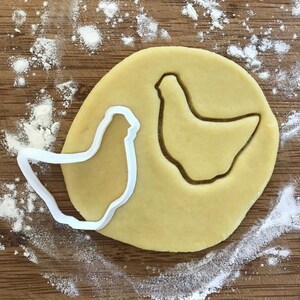 May include: A white plastic chicken-shaped cookie cutter next to a cutout in a circle of raw dough. The dough is on a wooden surface dusted with flour. The cookie cutter is used to shape the dough into a chicken.