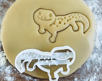 May include: A white plastic cookie cutter in the shape of a gecko lizard. The cutter is on top of a round, light brown cookie dough.