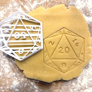May include: A white plastic cookie cutter in the shape of a 20-sided die, next to a cut-out of the same shape in raw cookie dough. The die shape has numbers and lines. The dough is on a wooden surface dusted with flour.