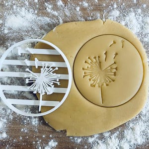 May include: A white plastic dandelion cookie cutter next to a circle of pressed dough with a dandelion imprint. The dough is light brown, and the cutter has a striped design. Flour is sprinkled around the dough.