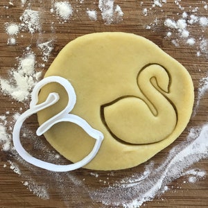 May include: A white plastic cookie cutter in the shape of a swan and an ampersand on top of a round, light brown cookie dough.