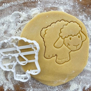Op de afbeelding: Een witte plastic koekjesvormer in de vorm van een schaap, met een gedetailleerd gezicht en wol, ligt op een rond stuk koekdeeg.