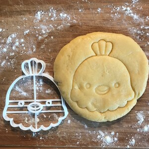 Puede incluir: Un cortador de galletas de pl&aacute;stico blanco con forma de pollito de dibujos animados con una corona en la cabeza, junto a una masa de galletas redonda cortada con el cortador.