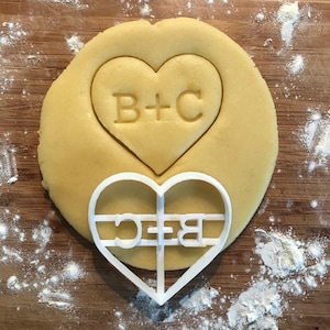 May include: A white heart-shaped cookie cutter with the letters "B+C" inside. The cutter is on top of a round, light brown cookie dough with the same letters imprinted on it.