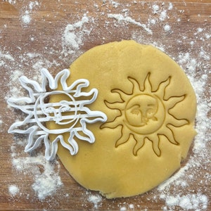 Pode incluir: Um cortador de biscoitos de plástico branco em forma de sol com um rosto e um padrão listrado. O cortador de biscoitos está em cima de uma peça redonda de massa de biscoitos não cozida com uma impressão de cortador de biscoitos em forma de rosto de sol.