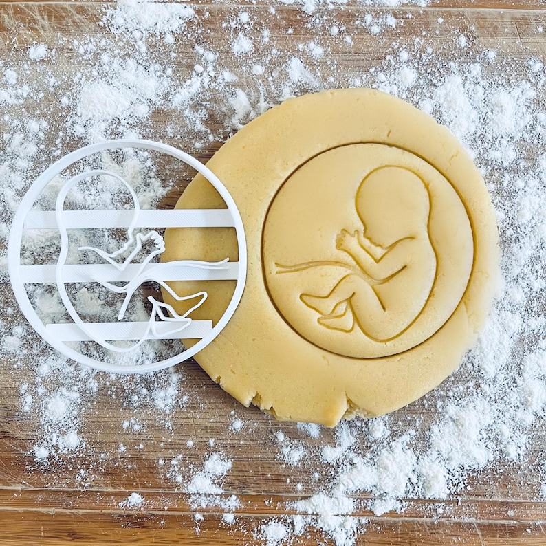 K&ouml;nnte beinhalten: Ein wei&szlig;er Plastik-Ausstecher in Form einer schwangeren Frau mit einem Baby im Bauch. Der Ausstecher liegt auf einer mit wei&szlig;em Mehl bedeckten Holzoberfl&auml;che.