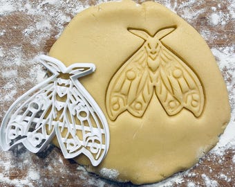 May include: A white plastic cookie cutter in the shape of a moth, with intricate details, is shown on top of a round piece of unbaked cookie dough. The cookie cutter is slightly offset to the left of the dough, and the moth design is visible in the dough.