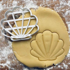 Könnte beinhalten: Ein weißer Ausstecher in Muschelform neben einem ausgerollten Teigstück mit einem Muschelabdruck. Der Teig ist hellgelb, und der Ausstecher liegt darauf. Mehl ist leicht auf der Holzoberfläche verstreut.