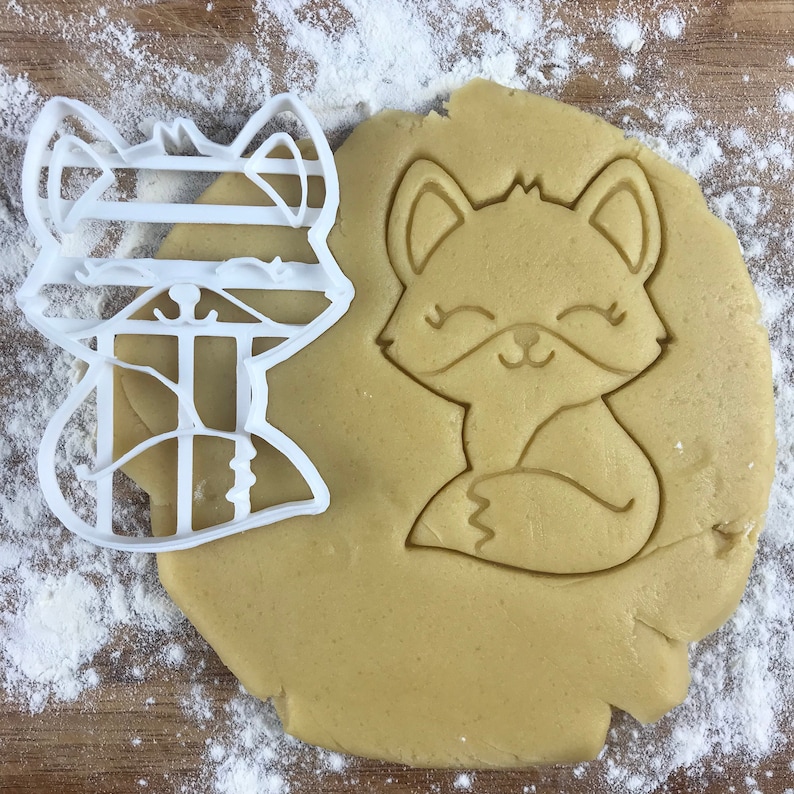 Puede incluir: Un cortador de galletas de pl&aacute;stico blanco en forma de zorro con un patr&oacute;n rayado en su cuerpo. El cortador est&aacute; al lado de un recorte de masa para galletas de un zorro con una cara sonriente.