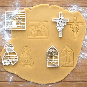 May include: A collection of white plastic cookie cutters on a sheet of dough. The cutters include shapes of crosses, a tomb, Jesus, and stained glass windows. The dough is light brown, and there is flour sprinkled around the edges.