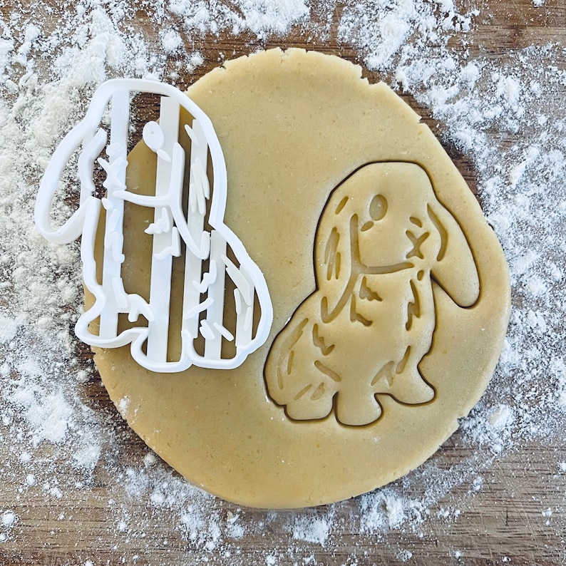 May include: A cookie cutter set featuring two animal designs: a striped rabbit and a fluffy bunny. The cutters are made of white plastic and are shown on a brown surface covered in white flour.