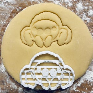 Op de afbeelding: Een witte plastic koekjesvormer in de vorm van een cartoon-spin. De vormer ligt op een rond koekdeeg. De spin heeft acht poten, twee grote ogen en een gestreept patroon op zijn lichaam.