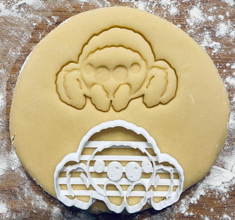 Op de afbeelding: Een witte plastic koekjesvormer in de vorm van een cartoon-spin. De vormer ligt op een rond koekdeeg. De spin heeft acht poten, twee grote ogen en een gestreept patroon op zijn lichaam.