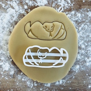 Puede incluir: Una masa redonda de color marrón claro con un cortador de galletas en forma de serpiente. El cortador es blanco y tiene un diseño de serpiente. La masa tiene una forma de serpiente en relieve. Harina espolvoreada alrededor de la masa sobre una superficie de madera.