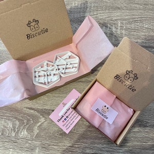 May include: A brown cardboard box with a pink tissue paper lining. The box contains two white plastic cookie cutters shaped like houses with a striped pattern. The box has a logo that says "Biscutie" with a cartoon deer. A thank you card with the same logo is visible in the foreground.