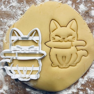 May include: A white plastic cookie cutter in the shape of a cat holding a knife. The cutter is on top of a round, light brown cookie dough.